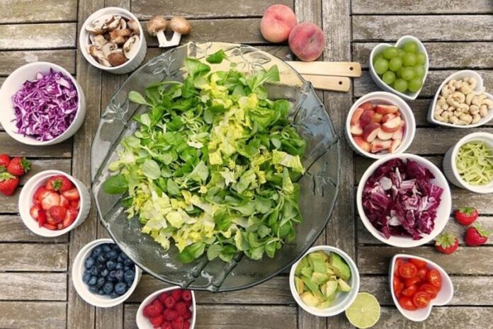 A large bowl of fresh mixed greens surrounded by bowls of colorful fruits, vegetables, and nuts on a wooden table.