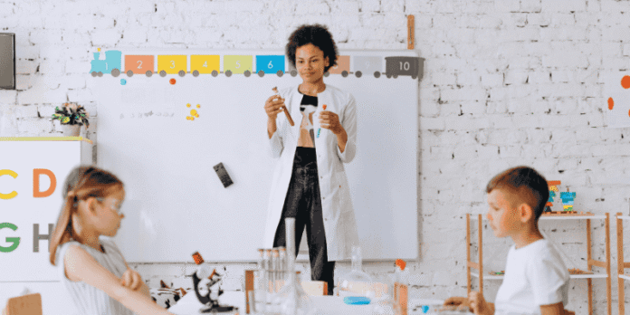 An early childhood educators in a lab coat demonstrates a science experiment to two children in a classroom. An early childhood educators in a lab coat demonstrates a science experiment to two children in a classroom.