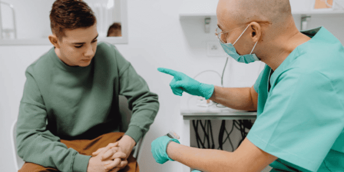 A healthcare professional wearing gloves and a mask talks to a teenage boy, providing medical guidance in a clinic setting.