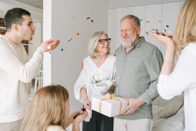 Elderly couple receiving gifts and flowers while family members throw confetti in celebration.