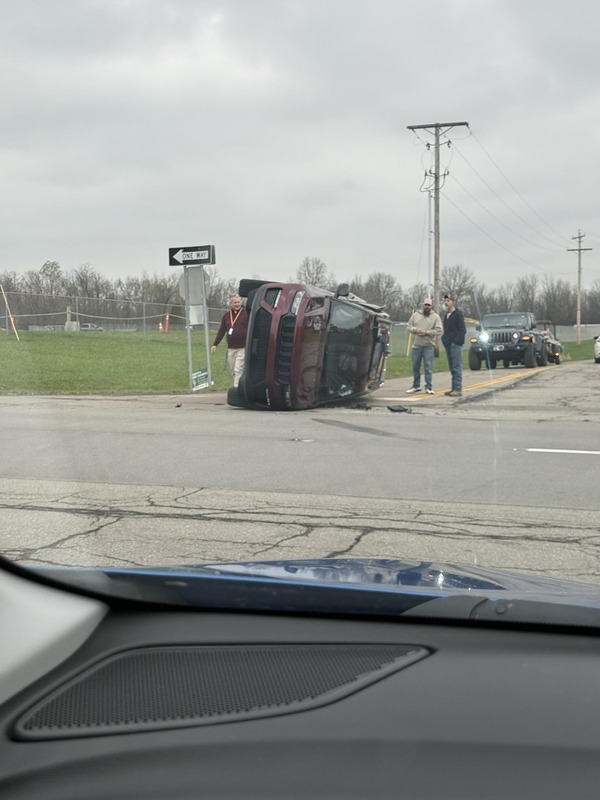 Maroon SUV accident with car flipped on its side, three men standing nearby, and another vehicle behind under cloudy skies.