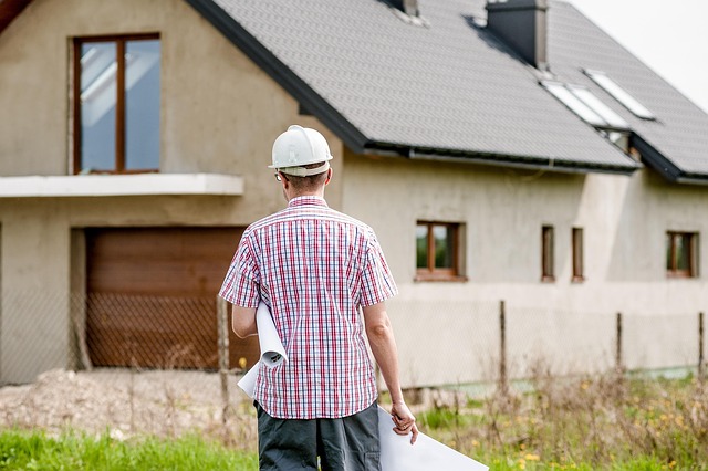 A man in a hard hat holding blueprints while inspecting a modern house under construction.