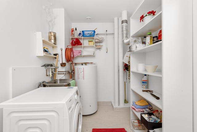 A laundry room with a water heater, washing machine, sink, and shelves filled with cleaning supplies and household items.