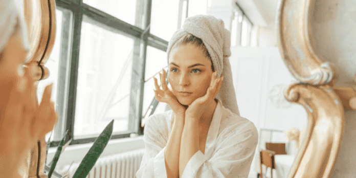 Woman with Towel Wrapped Hair Looking in Mirror A young woman with a towel wrapped around her head looks at herself in the mirror while gently touching her face, wearing a white robe.