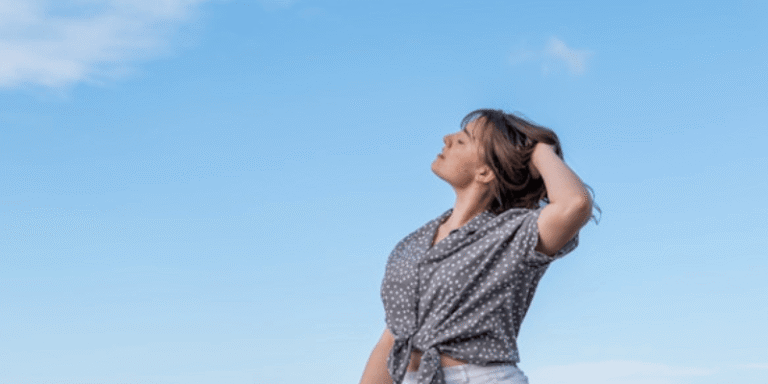 A woman in a gray polka dot shirt and white shorts stands outdoors with her hand in her hair, looking up at the clear blue sky, symbolizing Reinventing Yourself.