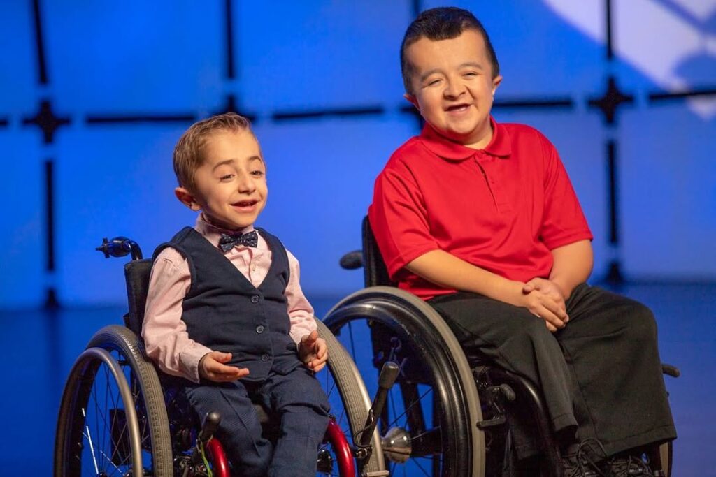 Alec and Kaleb sitting in wheelchairs, smiling on stage with a blue background.