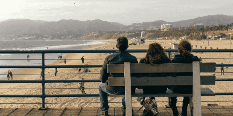 A family seated on a pier bench overlooking a sandy beach and coastal mountains, representing the beauty and lifestyle of California.