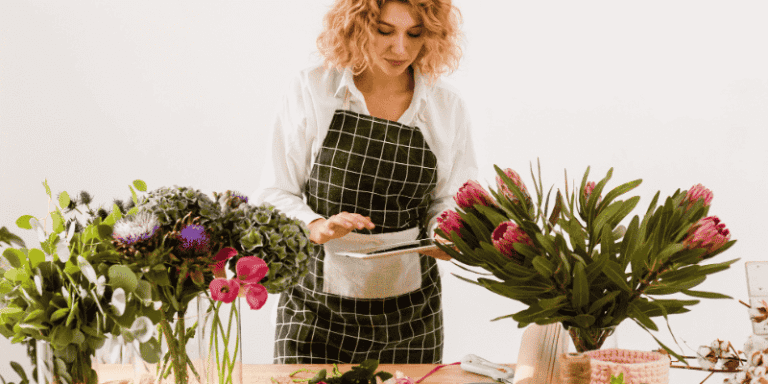 A female florist wearing an apron arranges vibrant flowers on a wooden table, using a tablet to organize her floral designs in a bright studio.