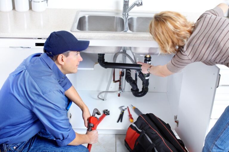 A professional plumber fixing a kitchen sink while a homeowner looks on, showing expert plumbing service and home maintenance.