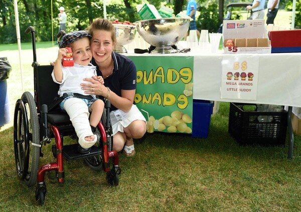 Kaleb-Wolf smiling with his mom Marie-Hélène Kaleb-Wolf sits in his wheelchair beside his mom Marie-Hélène in an outdoor setting, both smiling.