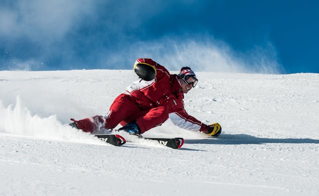 A male skier in a red and white ski suit is shown mid-action, carving a very deep turn on a snowy slope.