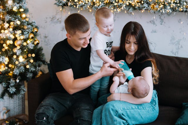A happy family sitting on a couch near a decorated Christmas tree, with parents and an older child helping to feed a baby with a bottle.