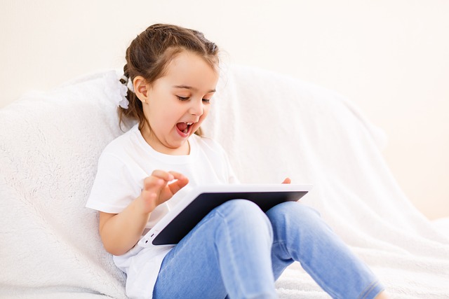 Child Engaged with Digital Device A kid girl smiling and enjoying while using a tablet on a couch, wearing a white t-shirt and blue jeans.