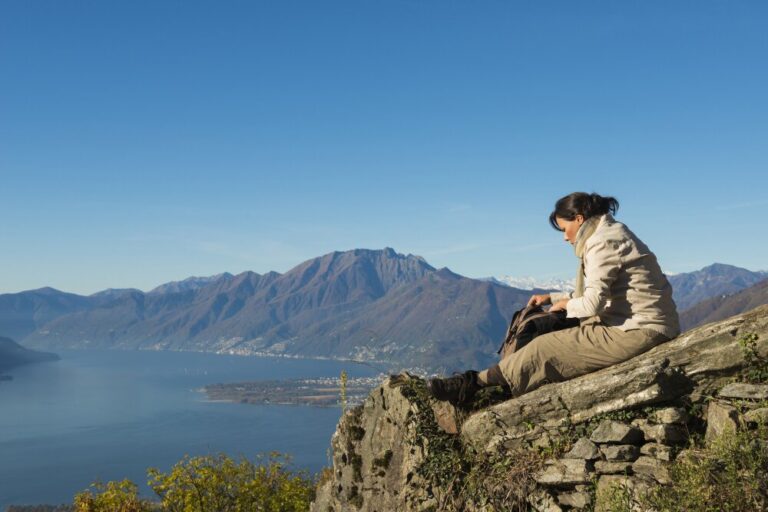 A woman sitting on a rocky ledge overlooking mountains and a lake, enjoying a quiet moment on a mountain vacation