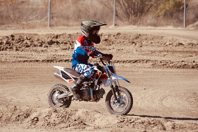 A young rider on a dirt bike, wearing full motocross gear including a helmet, riding on a dirt track. Kid Riding Dirt Bike with Helmet in Protective Gear