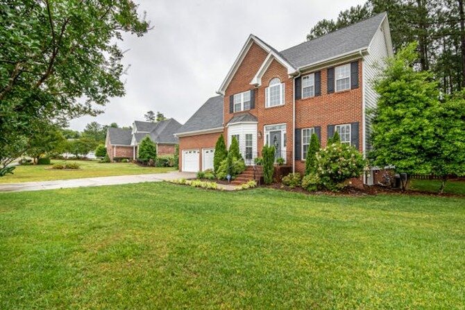 A two-story brick and siding suburban house with a neatly maintained front lawn, surrounded by trees and neighboring homes.