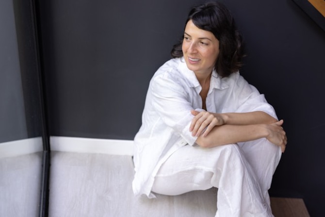 calm woman relaxing indoors while naturally coping with menopause symptoms at home. A calm woman dressed in white sits comfortably on the floor indoors, representing emotional balance and self-care while experiencing menopause symptoms.