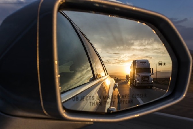 Car Side Mirror Reflecting Semi Truck on Sunset Highway for Safe Driving A car side mirror reflects a semi truck traveling on a sunset highway, highlighting road awareness and safe driving practices around large trucks.