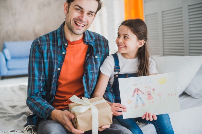 Memorable Gift Ideas to Celebrate Dad in 2026 Father smiling while holding a wrapped gift beside his daughter, who is showing a handmade drawing, representing thoughtful and meaningful gifts for dads.